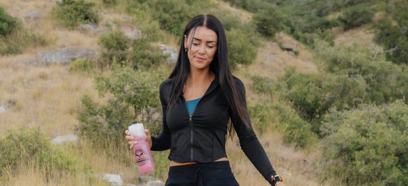 Woman walking on a trail in a natural setting holding a pink water bottle.