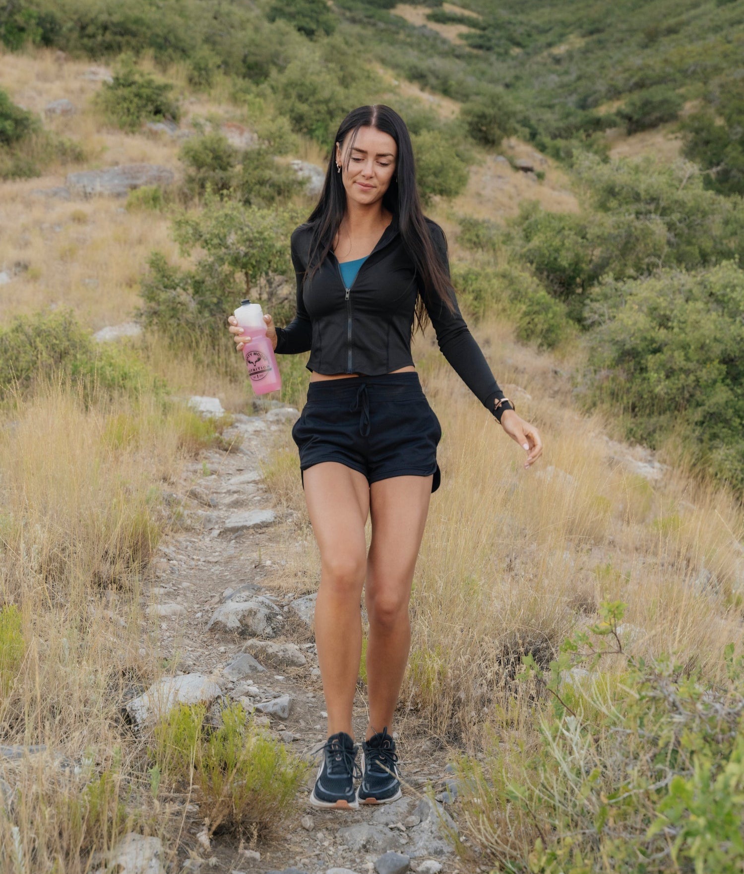 Woman walking on a trail in a natural setting holding a pink water bottle.